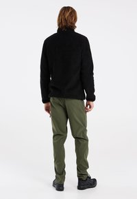 Zwarte fleecejack met hoge kraag, textuurstof, gecombineerd met olijfgroene lichte broek en zwarte sportschoenen.