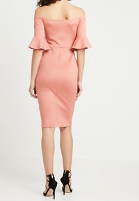 Společenské šaty bez ramínek, těsně přiléhající v růžově růžové barvě s rozšířenými rukávky. Hladká, elastická látka s délkou po kolena. Nosené s černými sandály na vysokém podpatku.