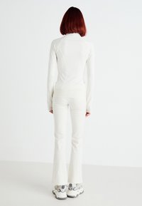 Top bianco aderente a maniche lunghe con colletto alto e dettagli cuciti; abbinato a pantaloni a zampa di elefante dello stesso colore e sneakers bianche.