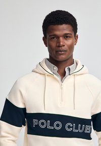 Crème hoodie met een ritssluiting in de hals, marineblauwe streep met "POLO CLUB" logo en groene accentpiping. Gelaagd over een gestreepte overhemden.