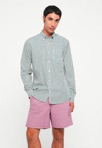 Stribet skjorte i grøn og hvid, med lange ærmer og en brystlomme, parret med rosa shorts. Ensfarvet baggrund.