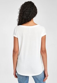Hvid T-shirt med korte, rullede ærmer og rundet ryg, lavet af et let, blødt stof; parret med blå jeans, set fra ryggen.