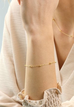 Gold chain bracelet featuring small round beads at intervals, worn on a light skin tone wrist. Soft, textured fabric visible in the background.