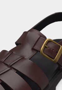 Καφέ δερμάτινο σανδάλι με πολλαπλές λουρίδες, αγκράφα σε χρυσαφί απόχρωση στη λουρίδα του αστραγάλου και μαύρη σόλα.