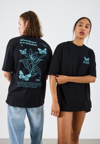 Sorte bomulds t-shirts har teal sommerfugle og blomster grafik på bagsiden, sammen med tekst. Løs pasform; korte ærmer. Enkel foran.