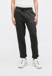 Tmavě šedé tepláky s elastickým pasem a šňůrkou, s fialovým logem "REEBOK". Kousek je stylizován s bílými teniskami.