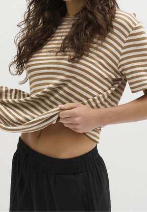 Kobieta podnosi brązowo-białą koszulę w paski, odsłaniając brzuch, nosząc czarne spodnie z elastyczną talią na białym tle.