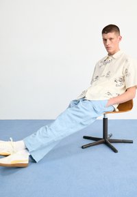 Korte mouwen crèmekleurig shirt met zwarte grafische prints, gecombineerd met lichtblauwe broek. Model zit op een houten stoel tegen een eenvoudige achtergrond.