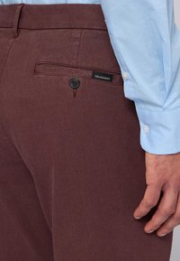 Burgundské kalhoty vyrobené z texturované tkaniny, s zadní kapsou na knoflík a značkovou visačkou. Košile je světle modrá.