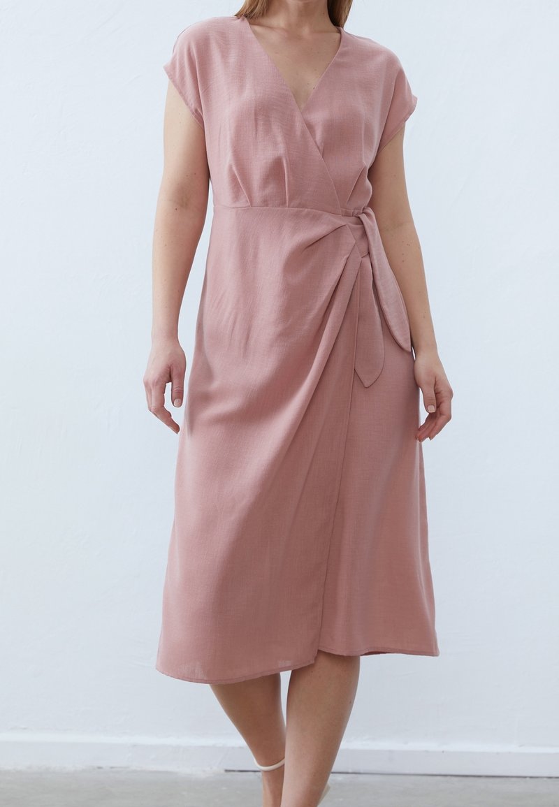 Femme portant une robe portefeuille rose poudré mi-longue à manches courtes, nouée à la taille, debout devant un fond clair uni.