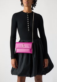 Rosa imitationsläder crossbody-väska med en silverbi-accent, med en vikbar klaff och en silverkedjerem, uppvisad mot en svart klänning.