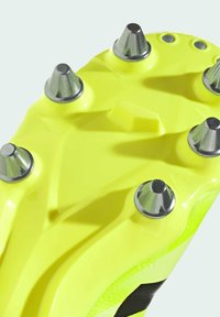 Semelle de chaussure de football jaune avec des crampons métalliques, présentant une surface texturée et un design aérodynamique. Des motifs géométriques notables sont présents.