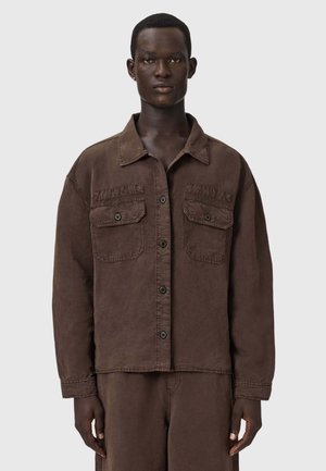 Man draagt een losse bruine jas met knopen en twee borstzakken, gecombineerd met bijpassende bruine broek, staand tegen een effen grijze achtergrond.