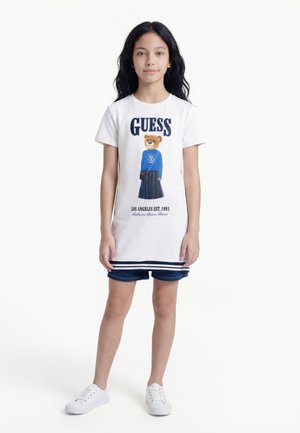 Dievča s dlhými tmavými vlasmi, oblečené v bielych šatách s grafikou medveďa a textom "GUESS", rifľových šortkách a bielych teniskách stojí na bielom pozadí.