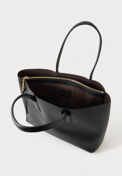 Sac fourre-tout en cuir noir avec deux poignées, ouverture en haut montrant l'intérieur marron foncé et une poche zippée dorée à l'intérieur.