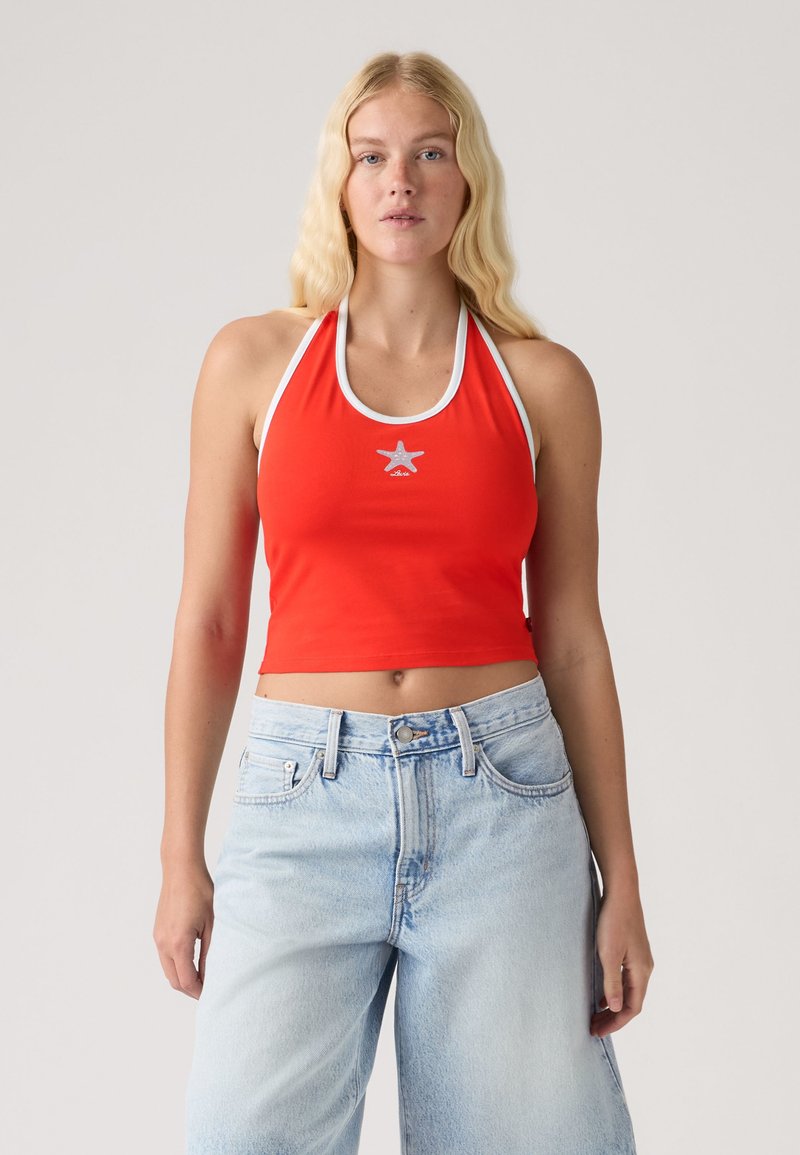 Ξανθιά γυναίκα που φοράει κόκκινο halter crop top με λευκή επένδυση και λογότυπο αστέρι, συνδυασμένο με ανοιχτό μπλε τζιν με ψηλή μέση.