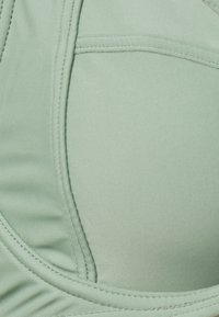 Sports bra v mint zeleni barvi, ki se ponaša z gladkim, raztegljivim materialom, obliko, ki se prilega telesu, in diskretnimi šivi ob robovih.