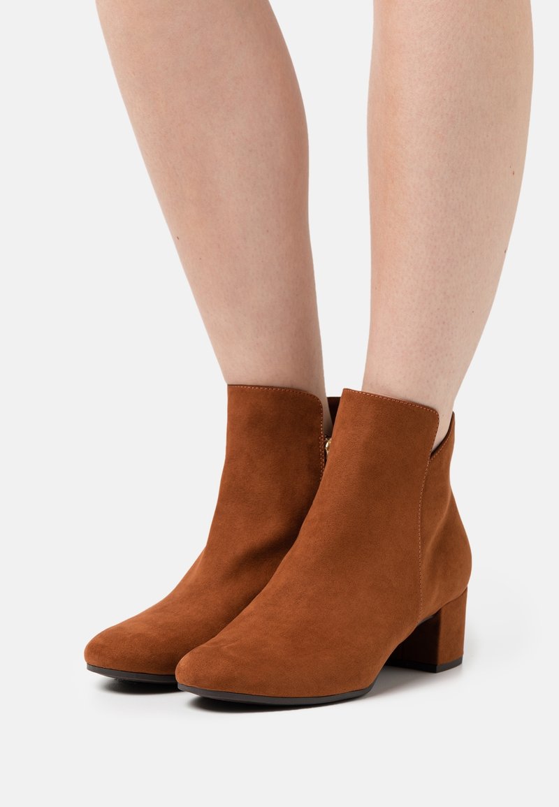 Paire de bottines marron en daim avec talons blocs portées sur jambes nues devant un fond uni.