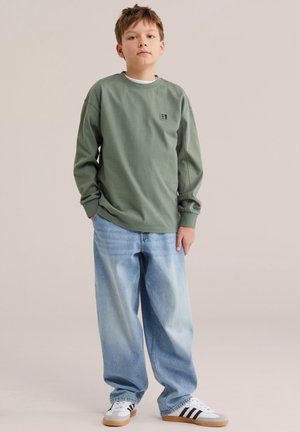 Poika, joka käyttää vihreää pitkähihaista paitaa, vaaleansiniset leveälahkeiset farkut ja valkoiset tennarit, seisoo yksinkertaisen beige taustan edessä.