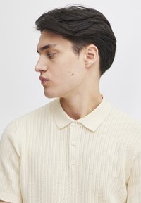Sweter polo w jasnej kremowej kolorystyce, z teksturowanym wzorem, krótkimi rękawami i kołnierzykiem, z trzema guzikami.