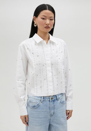 White cropped button-up shirt with vertical pintuck details, embellished with small silver studs, paired with light blue denim jeans.