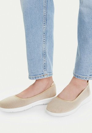 Pies con zapatos beige de punto sin cordones con suelas blancas y jeans azules claros recortados sobre un fondo liso.