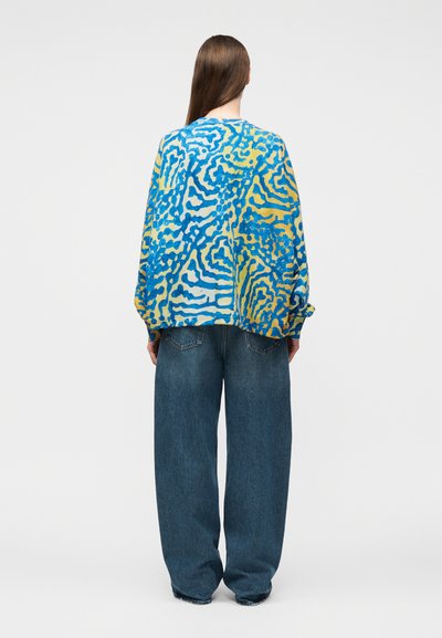 Άτομο με μακριά καστανά μαλλιά που φοράει ένα φαρδύ πουλόβερ μπλε και κίτρινο με σχέδιο και φαρδιά σκούρα μπλε τζιν, στεκόμενο με την πλάτη προς το μέρος μας.