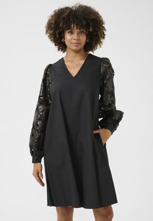 Abito nero con scollo a V e maniche corte di pizzo voluminose con motivi floreali. Include tasche laterali per funzionalità.