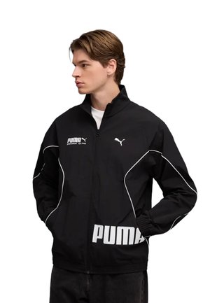 Giovane uomo che indossa una giacca nera Puma con bordi bianchi, loghi Puma sul petto e sulla manica, mani in tasca, guarda a sinistra.