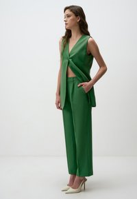 Débardeur vert sans manches avec un détail de bouton et un design cropped, associé à un pantalon droit assorti et des chaussures à talons hauts crème.