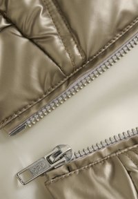 Tan nylon pufferjacka med krämfärgad foder, med dubbla metalldragkedjor och sömnadsdetaljer. Slät, glänsande yta.