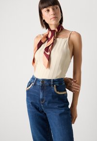 Top in cotone a costine beige con spalline sottili, abbinato a jeans a vita alta in denim blu con tasche decorate in oro e una sciarpa in seta stampata.