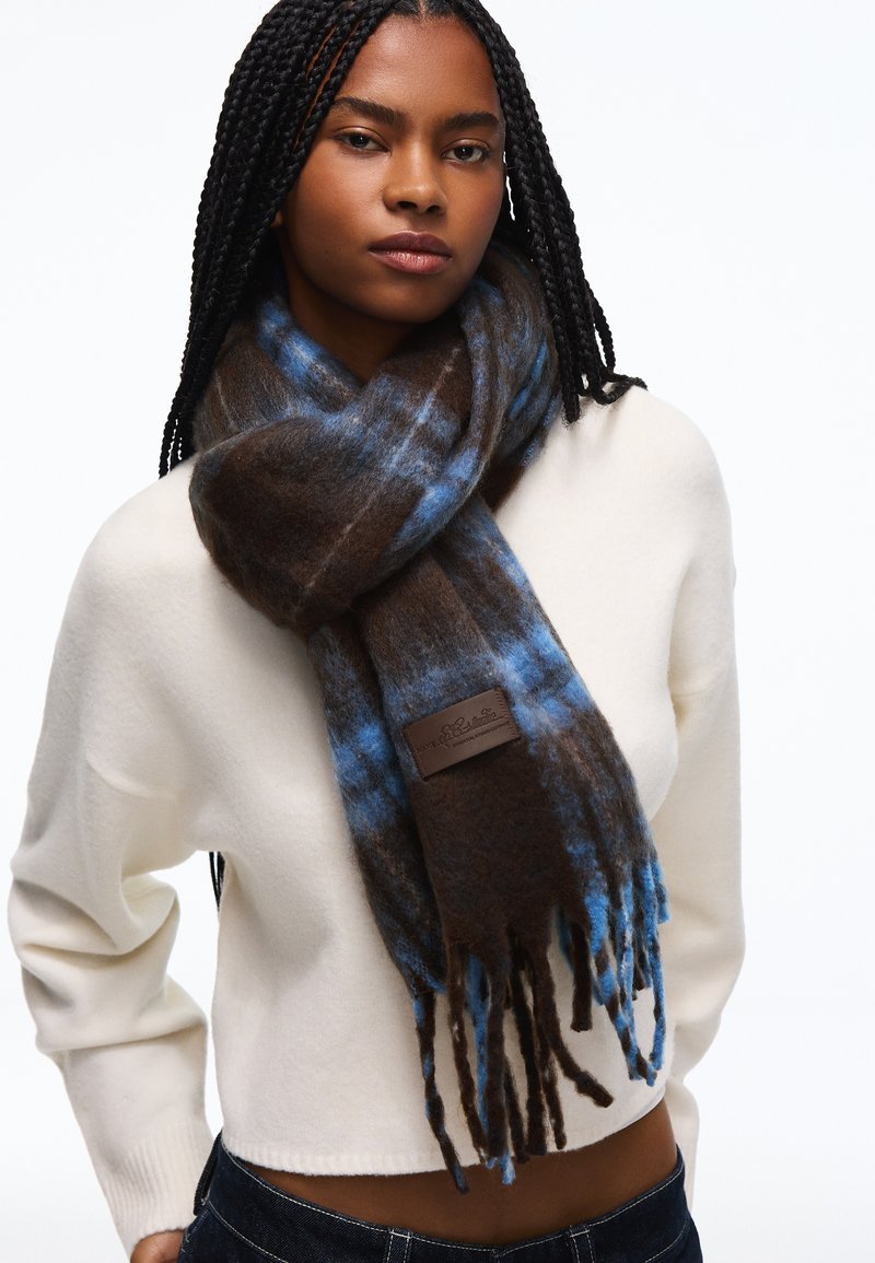 Femme aux longues tresses portant un pull blanc et une épaisse écharpe à carreaux marron et bleue avec des franges autour du cou.