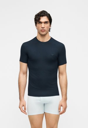 Muž v priliehavom čiernom tričku s krátkym rukávom a bielych boxerkách stojaci pred jednoduchým svetlým pozadím, otočený čelom vpred.