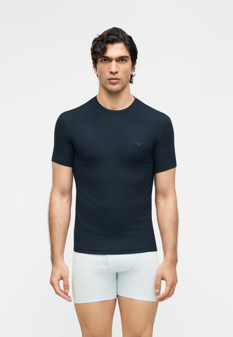 Άνδρας που φοράει εφαρμοστό μαύρο κοντομάνικο πουκάμισο και λευκά μπόξερ, στέκεται μπροστά σε απλό φωτεινό φόντο, κοιτώντας μπροστά.