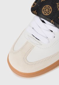 Sneaker met een witte leren bovenkant, tan suède accenten en een zwarte patroonriem met gouden ontwerpen, afgewerkt met een rubberen gumzool.