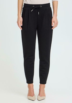 Femme portant un pantalon noir fuselé avec une taille à cordon et des plis, associée à des chaussures beige à bouts pointus, debout devant un fond blanc.