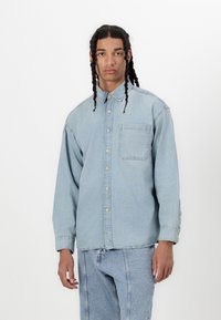 Cămașă din denim ușor albastru, cu nasturi, dotată cu un buzunar pe piept, mâneci lungi și croială lejeră. Textura ușor decolorată adaugă profunzime.
