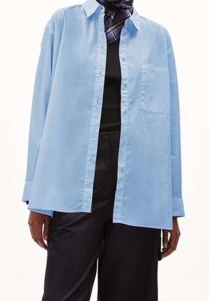 Lichtblauw overhemd met een relaxed fit, voorzien van een borstzak en lange mouwen, gecombineerd met een zwart topje en donkere broek.