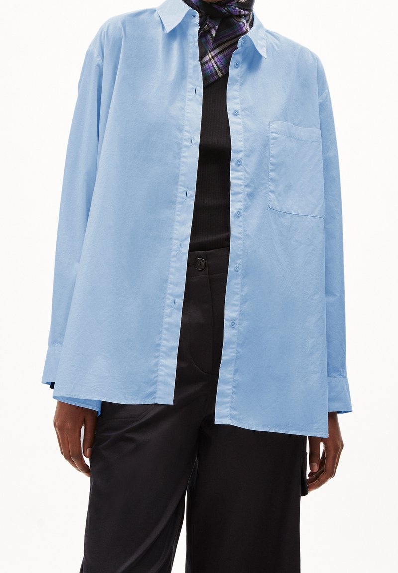 Light blue button-up shirt in a relaxed fit, featuring a chest pocket and long sleeves, paired with a black top and dark pants.