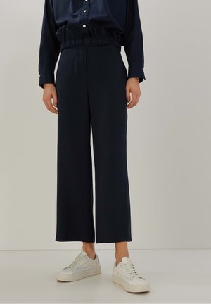 Personne portant un pantalon noir taille haute à jambes larges, une chemise bleu marine à manches longues et des baskets blanches, debout devant un fond uni.