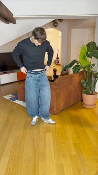 Široke plave traperice s laganim pranjem, uparene s crnom majicom dugih rukava. Srebrni sportski cipele; prostorija ima drveni pod i kauč.