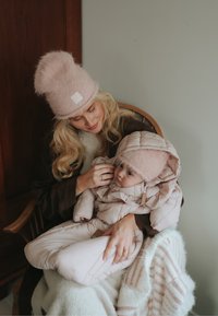 Ljusrosa fluffig mössa och matchande babymössa; kvinna i en mörkbrun kappa håller ett barn i en ljus överdragsdräkt i quiltat tyg på en trästol.