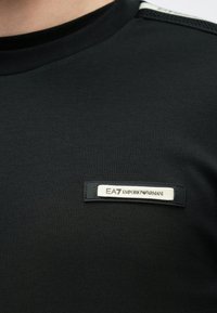 Melnas adītas auduma krekls ar baltu EA7 Emporio Armani logotipu uz krūškurvja pie apkakles.