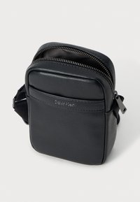 Bolsa transversal preta feita de material sintético texturizado. Apresenta um fecho de zip e detalhe do logótipo na frente, com uma alça ajustável.