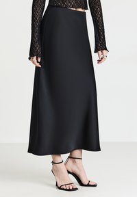 Donna che indossa una gonna midi di raso nera, una maglietta nera in pizzo con maniche lunghe, anelli d'argento e sandali con tacco alto e cinturino alla caviglia neri.