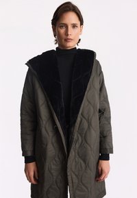 Pikowana oliwkowozielona kurtka z czarnym sztucznym futrem przy kołnierzu, o długim kroju, z zapięciem na guziki z przodu i bocznymi kieszeniami.