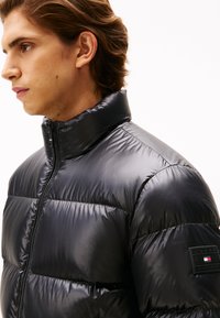 Black puffer jacket with a high collar, quilted texture, and smooth finish. Features a logo patch on the left sleeve and front zipper closure.