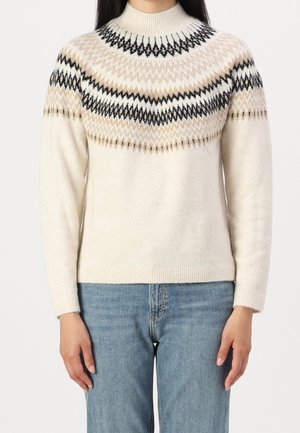Femme portant un pull en maille crème avec un motif géométrique noir et beige sur l'empiècement, associé à un jean bleu.