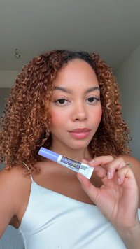 Une femme aux cheveux bouclés tient un tube d'anti-cernes Maybelline Superstay, portant un débardeur blanc devant un fond uni.